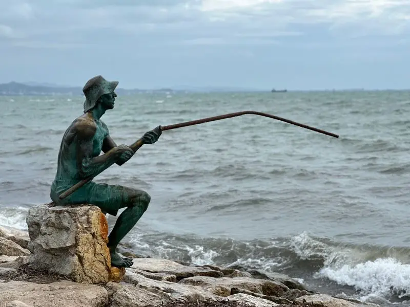 A bronze statue of a fisherman sits by the rocky seashore casting a fishing rod.