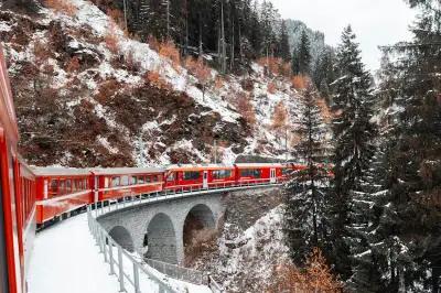 A vibrant red train crosses a snowy coniferous landscape on a scenic mountain bridge in winter.