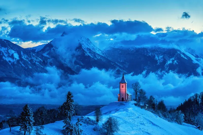 A serene winter scene of a church on a snowy hill with mountains and clouds during blue hour.