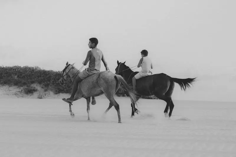Two boys ride horses across a sunny beach in Canoa Quebrada, Brazil, creating a dynamic scene of motion in black and white.