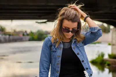 Fashionable woman in denim jacket with sunglasses stands under a bridge by the river.