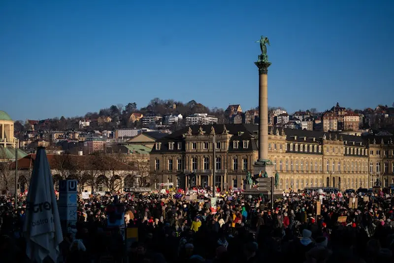 A large crowd gathers at Schlossplatz, Stuttgart, with historic buildings and landmarks in the background.