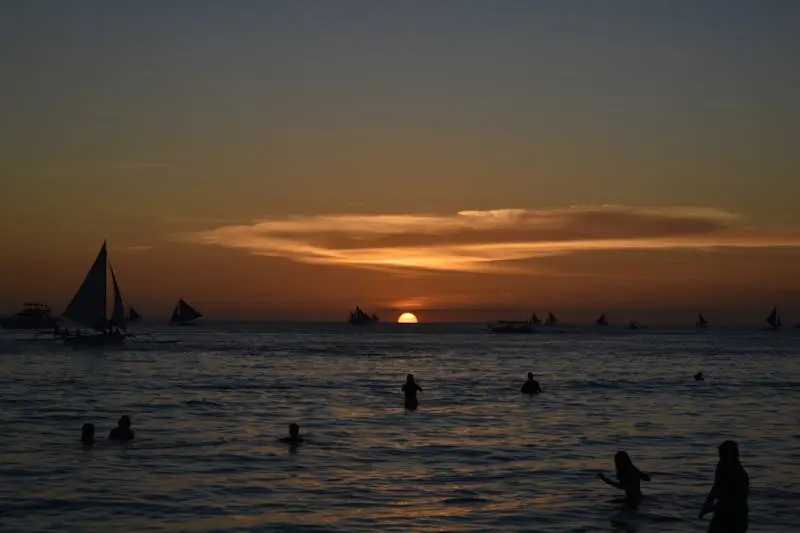 Stunning sunset silhouette on a tropical beach with boats and swimmers.