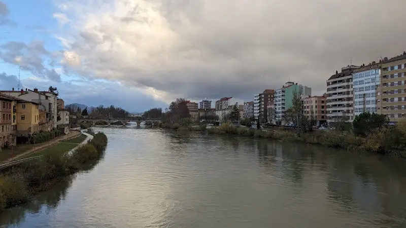 A tranquil river winds through a cityscape under a dramatic sky, featuring bridges and buildings.