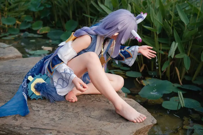 A person in elaborate cosplay costume sitting by a serene pond, touching the water.