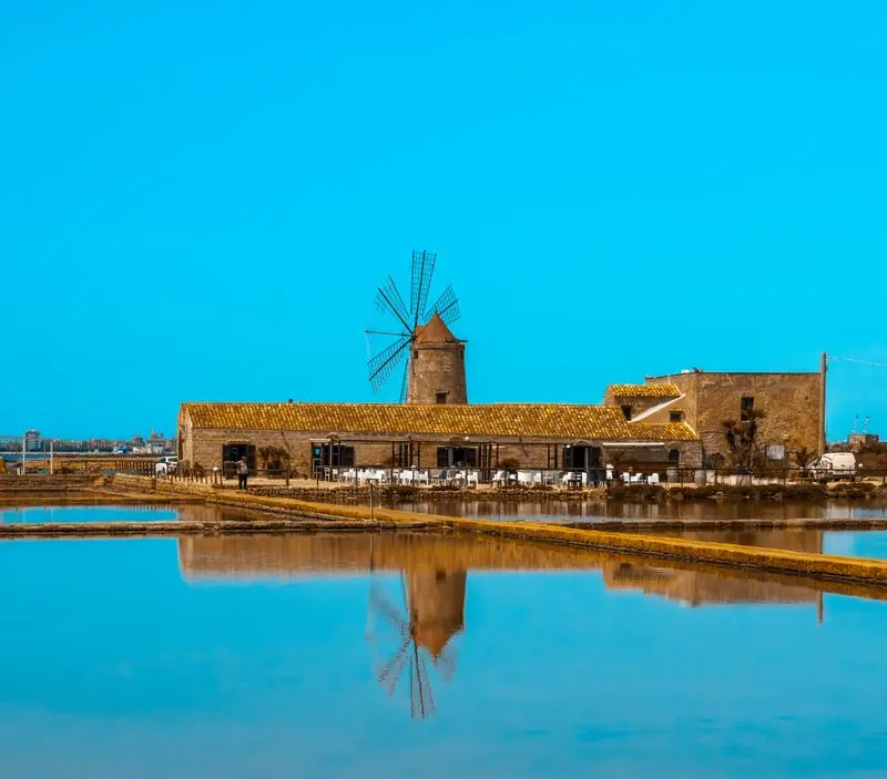 Stunning view of a historic windmill at Trapani Salt Works, Sicily, with mirror-like reflections.